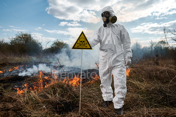 rotective radiation suit and gas mask near burning grass with smoke, holding yellow triangle with skull and crossbones warning sign.