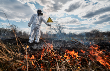 re. Man in protective suit and gas mask near burning grass with smoke, holding warning sign with skull and crossbones. Natural disaster concept.