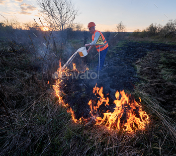 evening sky on background. Male firefighter pouring water on burning dry grass near yellow triangle with skull and crossbones warning sign.