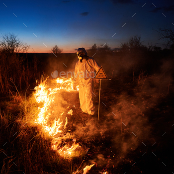 ght. Man in protective radiation suit and gas mask near burning grass with smoke and yellow triangle with skull and crossbones warning sign.