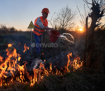 he evening. Male environmentalist holding bucket and pouring water on burning dry grass near yellow triangle with skull and crossbones warning sign.