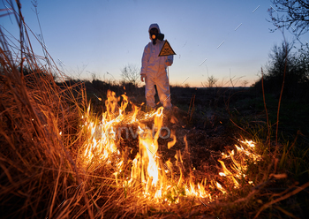 at night. Man in suit and gas mask near burning grass with smoke, holding warning sign with skull and crossbones. Natural disaster concept.