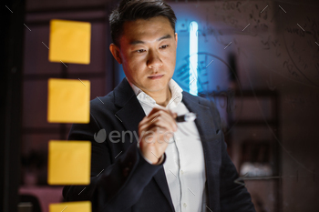 tasks in modern evening office. Close up of concentrated asian businessman in formal wear writing notes, charts and working tasks on glass wall.