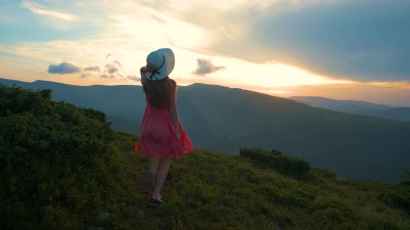 Young Woman in Red Dress Walking on Hillside Meadow on a Windy Evening in Summer Mountains