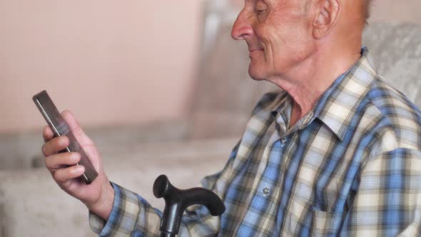 caucasian peppy pensioner with a smartphone and a walking stick sits on the couch at home.Smiling el