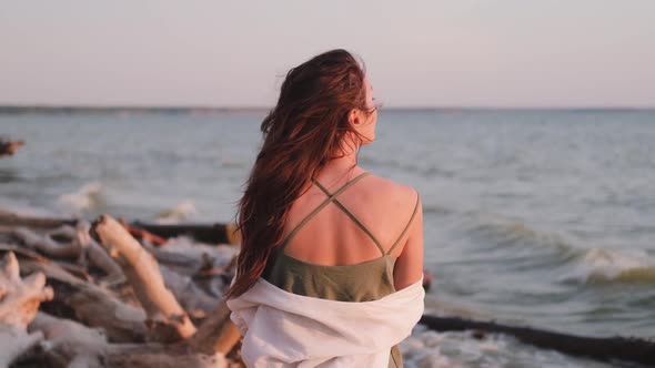 Young Woman Walks Along the Beach in a White Shirt on a Sunset Background. Silhouette of a Free Girl