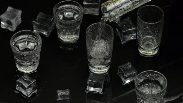Barman Pour Frozen Vodka From Bottle Into Shot Glass. Ice Cubes Against Dark Wet Black Background