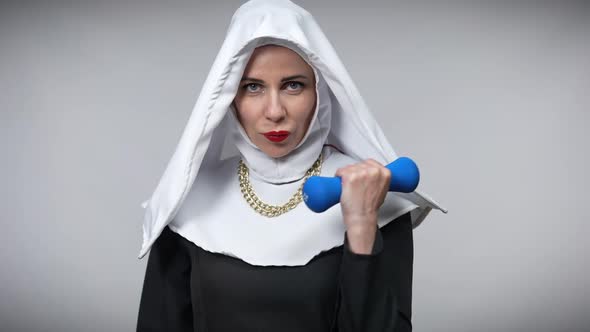 Confident Sportswoman in Nun Costume Lifting Dumbbell Looking at Camera Posing at Grey Background