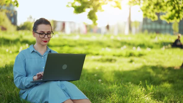 Busy Attractive Woman Working at the Laptop As Sitting on Grass in City Park