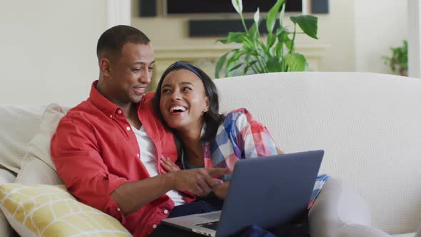 Happy biracial couple sitting on sofa, embracing, using laptop and laughing