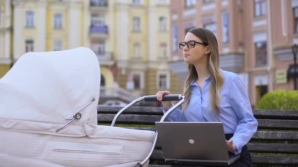 Multitasking Mother Swinging Baby Carriage and Working Laptop Sitting on Bench