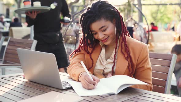 Young African American Woman Sitting in Outddor Cafe with Laptop and Making Notes in Notepad