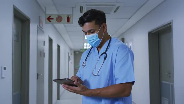 Mixed race male doctor wearing face mask standing in hospital corridor using tablet