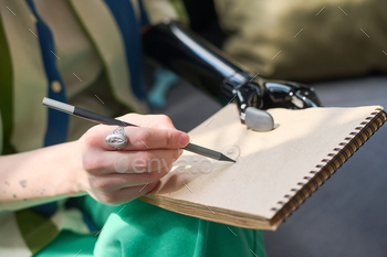 Hand of young woman with physical disability holding pencil over page of notepad