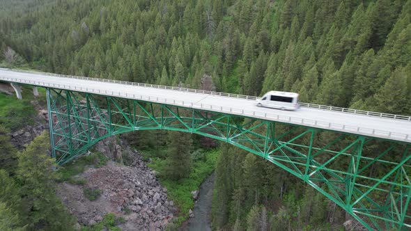 Drone watches as a van drives across a green painted steel bridge at the mouth of a pine covered can