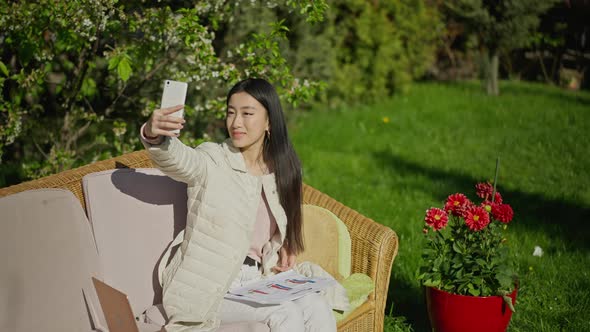 Happy Asian Millennial Woman Taking Selfie on Smartphone Sitting in Sunny Garden with Paperwork and