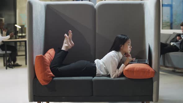 Asian Female Office Manager Lies in a Large Chair and Works at a Laptop Talking on a Conference Call