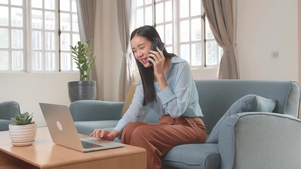 Asian Woman Sitting Sofa In Living Room While Speaking By Phone And Working On Laptop