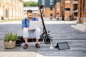 Man charges laptop with solar panels while sitting on electric scooter outdoors