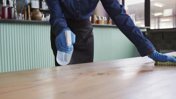 African american woman wearing gloves and apron disinfecting tables at cafe bar