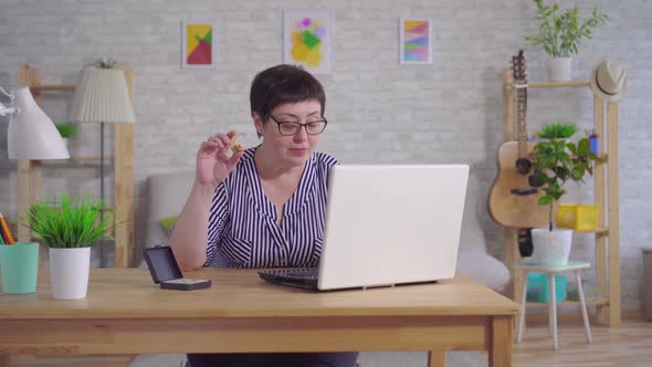 Smiling Woman with Glasses Sitting at a Table in the Living Room Holding a Hearing Aid Uses a Laptop