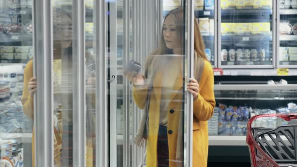 A Woman in a Fashionable Orange Coat Opens the Refrigerator in a Dairy Store