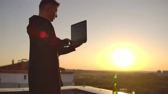 A Male Stockbroker Freelancer Stands on a Rooftop at Sunset with a Laptop and Types on a Keyboard