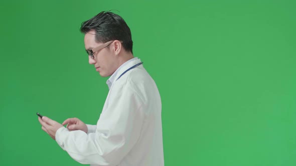 Side View Of Asian Doctor With Stethoscope Using Mobile Phone While Walking On Green Screen