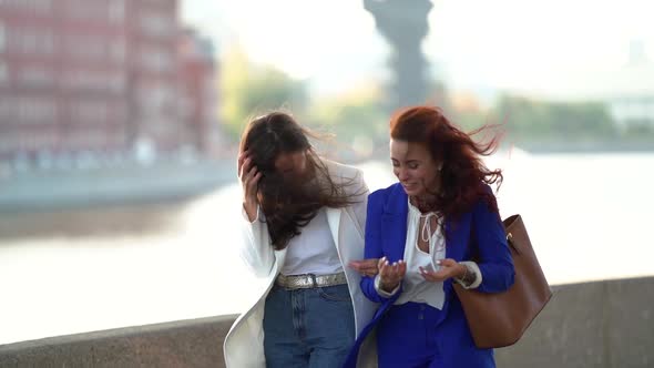 Two Business Women in Suits Walk Through the Spring City Past the River Together. Two Friends Talk