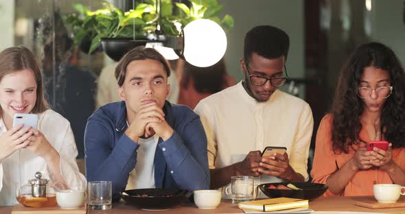 Young Bored Guy Sitting Between Gadget Addicted Friends with Smartphones in Cafe. Millennial People