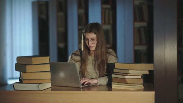 Girl Working with Laptop at the Desk with Books and Smiling in Library Late