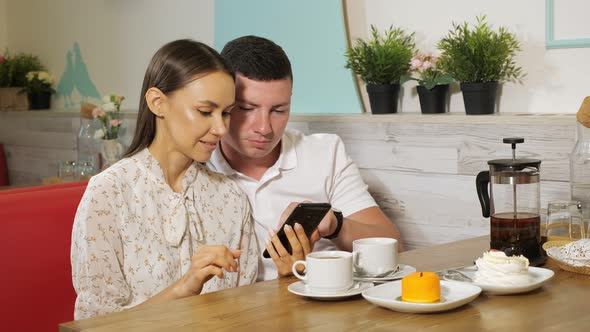 Girl and Guy Looks at Mobile Phone at Table in Sweet Shop