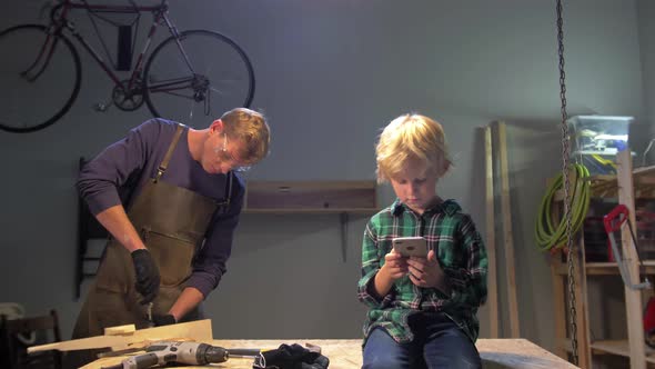 Boy Sits with Phone, Man Works in a Workshop