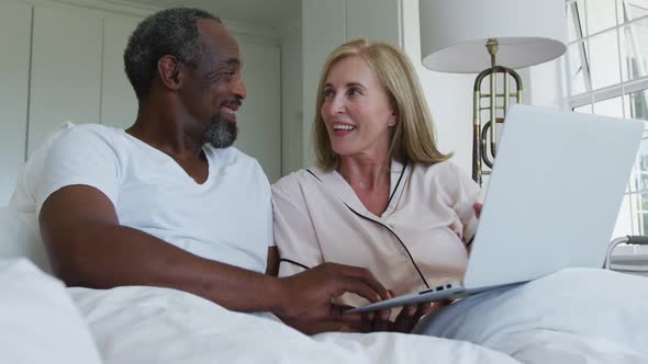 Diverse senior couple sitting in bed using laptop talking and laughing