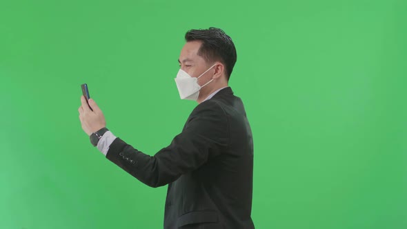 Asian Business Man Having Video Call On Phone While Walking On Green Screen, Wear Medical Mask