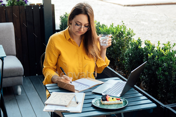 writes in a notebook and working with laptop in street cafe at summer day.
