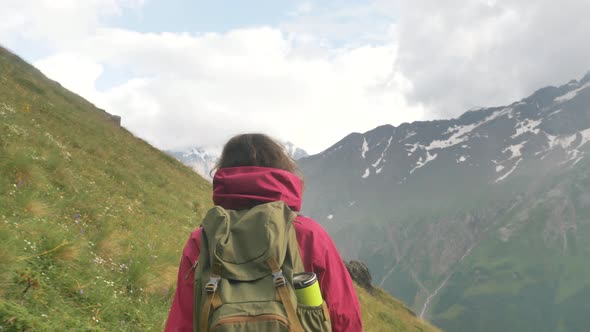 A Female Tourist Walks Along a Green Slope on a Mountain Path Against the Background of Snowcapped