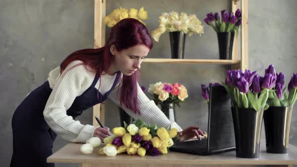 Florist in an Apron Working in Her Own Flower Shop Using a Laptop and Writing Down an Order for a