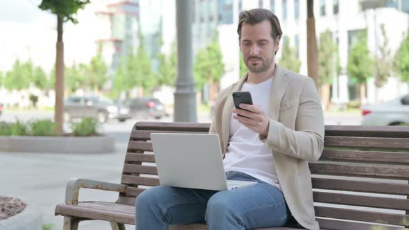 Young Man Using Smartphone and Laptop While Sitting on Bench