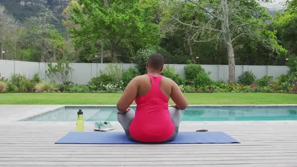 Happy african american plus size woman practicing yoga, stretching next to swimming pool in garden
