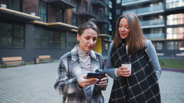 Two Happy Women Walking with Takeaway Coffee and Talking with Interest Among Themselves in the