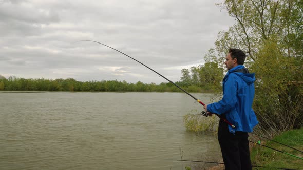 An angler in a blue jacket with a long fishing rod stands on the bank of the river