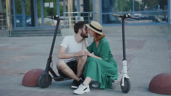 Young Lovely Couple Sitting Between Electric Scooters and Kisses Each Other