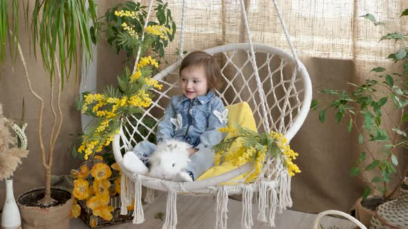 Little Girl is Riding in a Wicker Swing and Stroking a White Live Rabbit on a Festive Easter Day