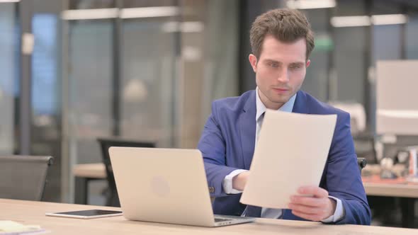 Businessman with Laptop Reading Documents in Office