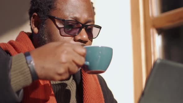 Pensive African man in eyeglasses looking at tablet and drinking coffee