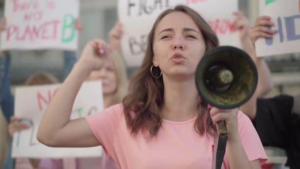 Young Female Eco-activist Yelling Through Megaphone with Crowd of People at the Background. Group of