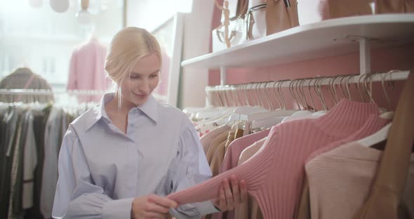 Young Attractive Blonde Woman Carefully Examines Clothes on a Rack or Hanger in a Shopping Center