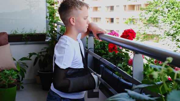 Sad Child with a Broken Arm in a Cast Stands on the Balcony and Waves to Someone