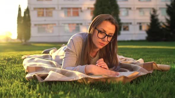 Girl in Glasses Reading Book Lying Down on a Blanket in the Park at Sunset